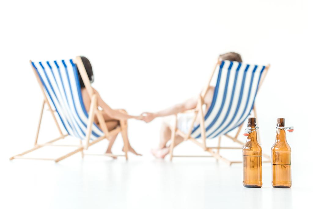 επιλεκτική εστίαση του ζευγάρι κρατώντας τα χέρια και να στηρίζεται σε καρέκλες παραλίας, με μπουκάλια μπίρας σε πρώτο πλάνο, απομονώνονται σε λευκό - Φωτογραφία, εικόνα