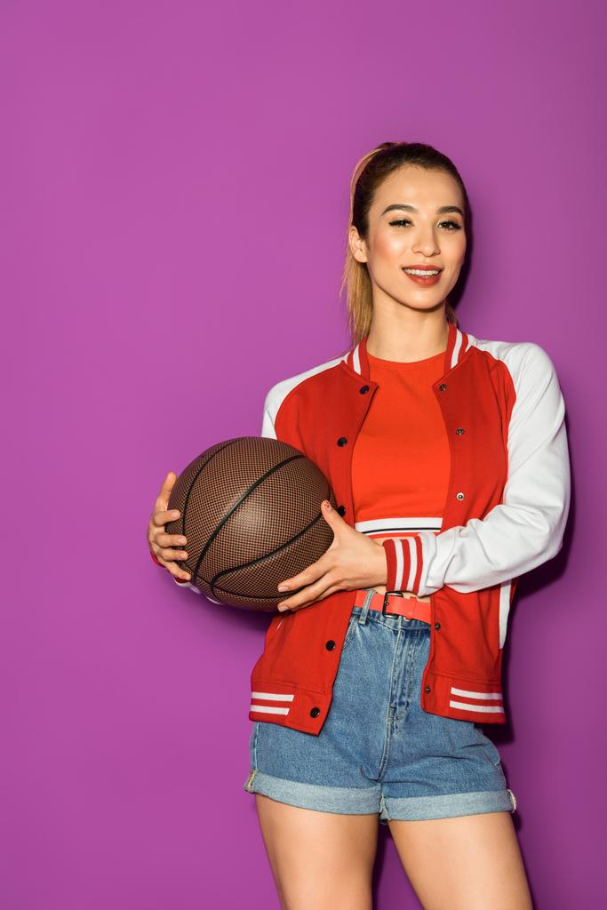 beautiful asian girl holding basketball ball and smiling at camera isolated on violet - Photo, Image