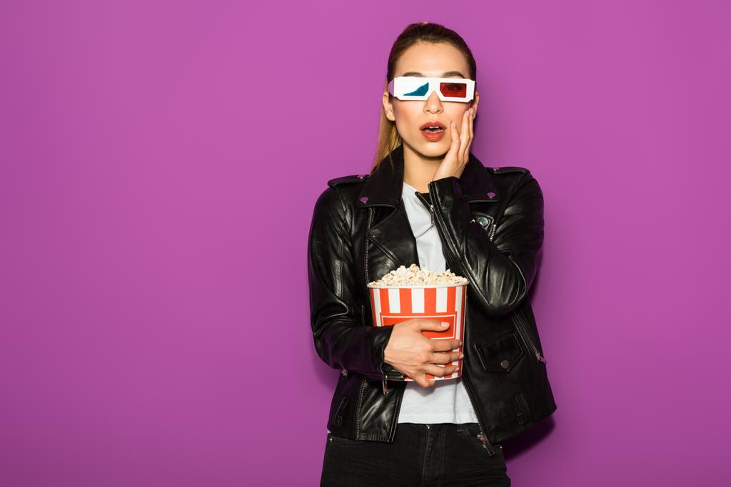 шокована молода жінка в 3d окулярах їсть попкорн і дивиться на камеру ізольовано на фіолетовому
  - Фото, зображення