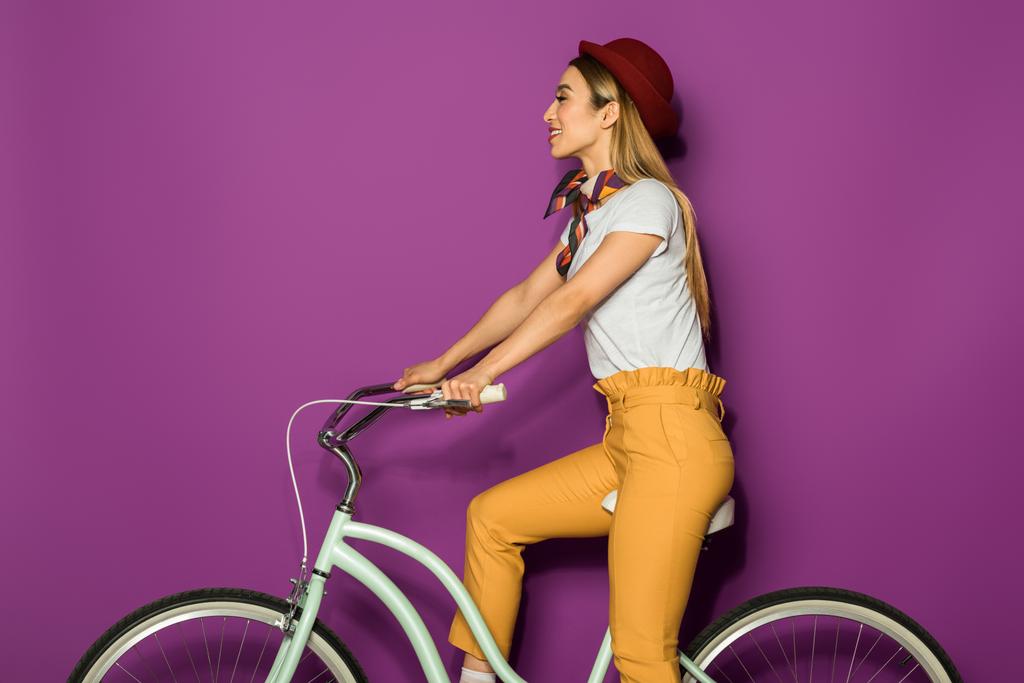 приваблива усміхнена стильна дівчина сидить на велосипеді і дивиться ізольовано на фіолетовому
 - Фото, зображення