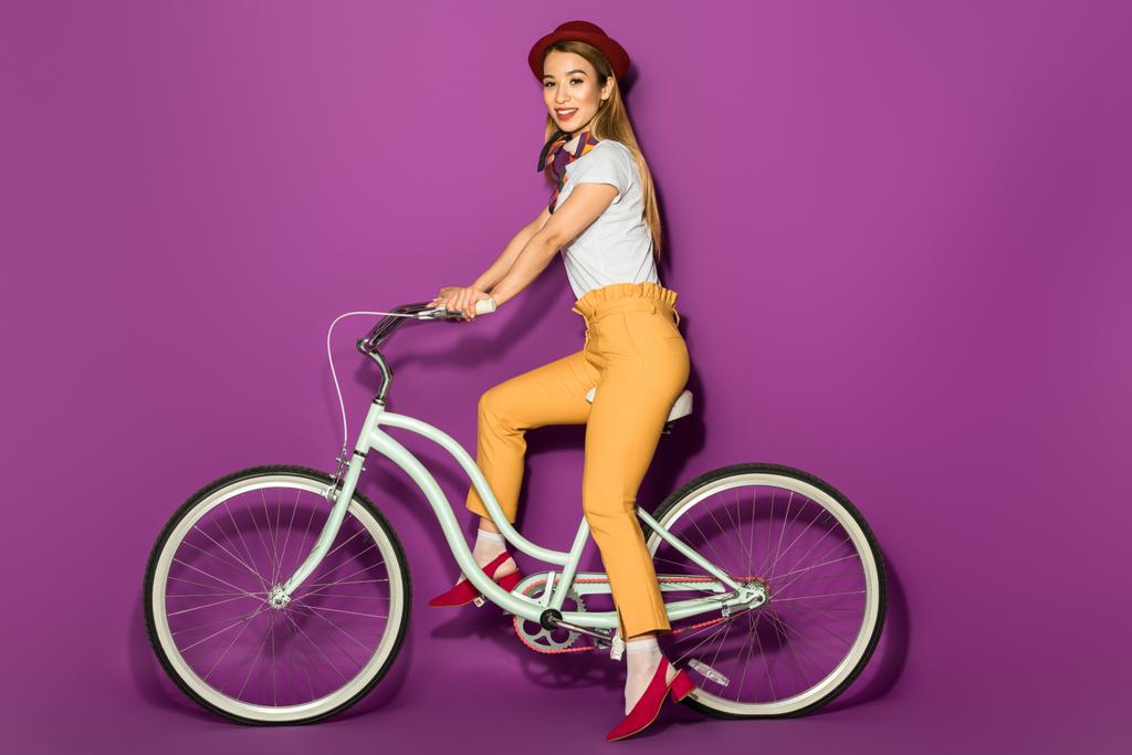 bonito elegante asiático menina equitação bicicleta e sorrindo para câmera isolado no violeta
 - Foto, Imagem