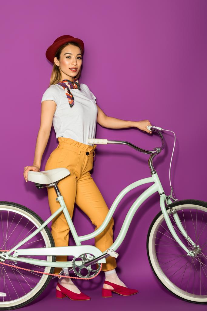 comprimento total de bela mulher asiática elegante de pé com bicicleta e sorrindo para a câmera isolada em violeta
  - Foto, Imagem