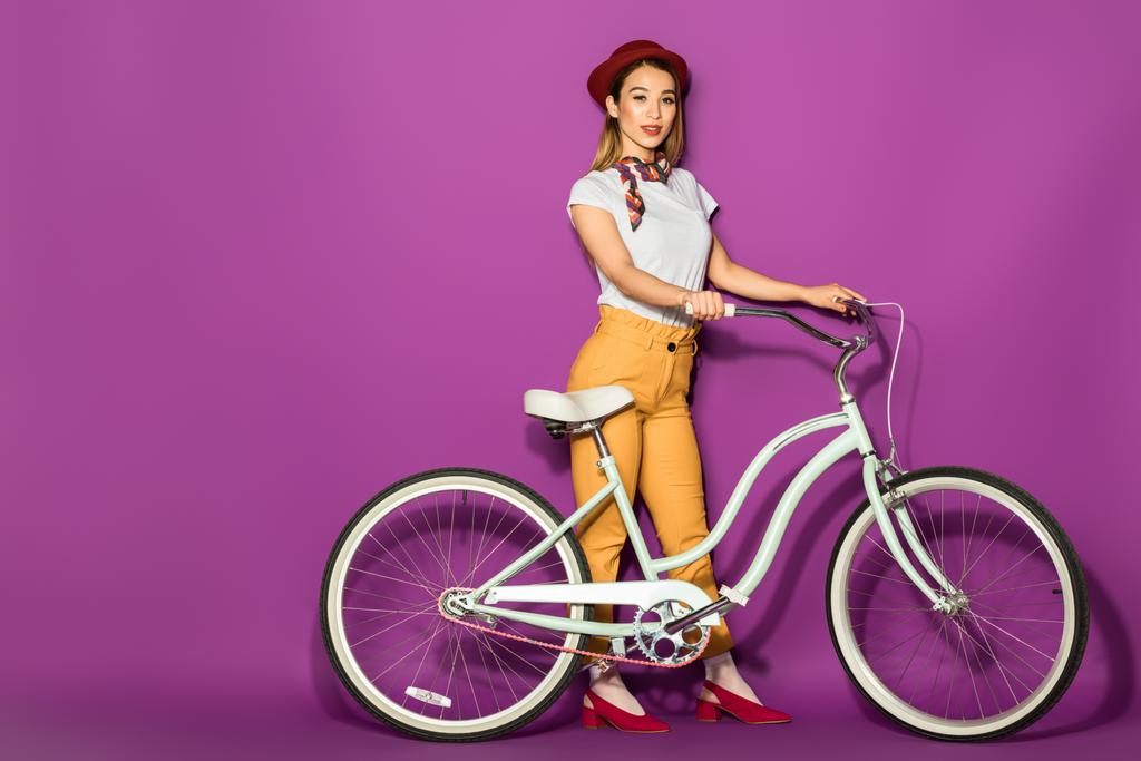 красивая стильная азиатская девушка стоит с велосипедом и улыбается на камеру изолированы на фиолетовый
  - Фото, изображение