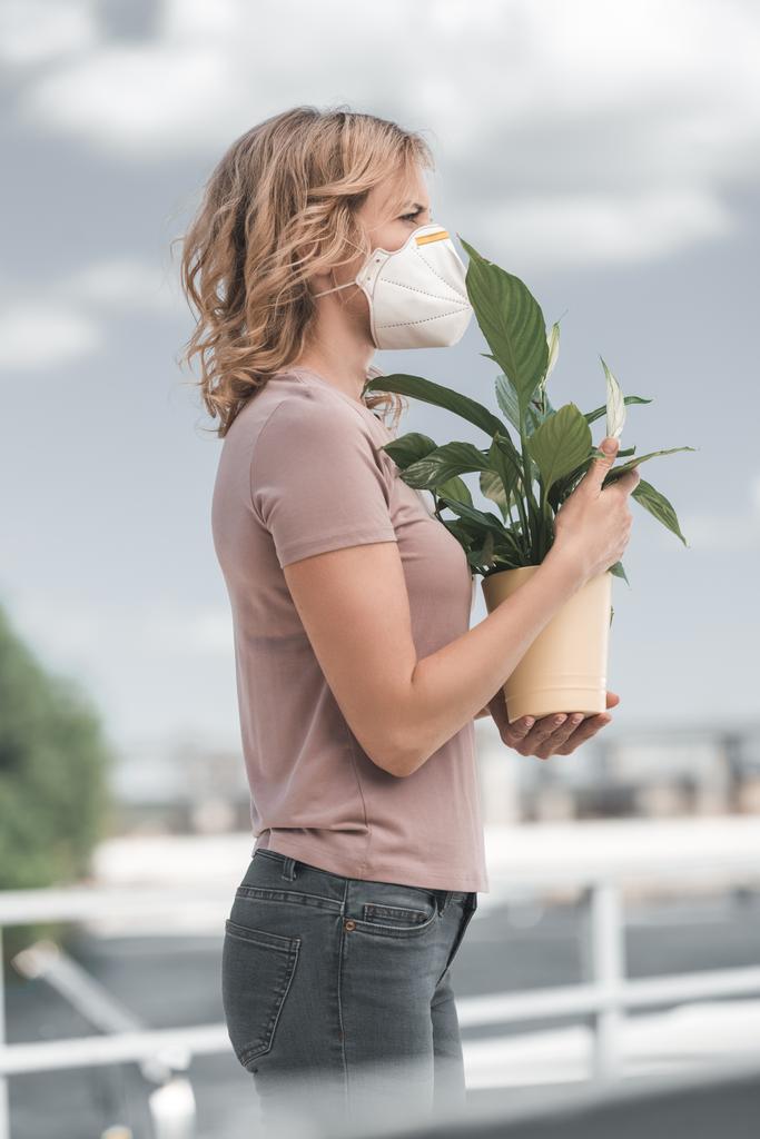 vista lateral da mulher na máscara protetora que prende a planta potted na ponte, conceito da poluição do ar
 - Foto, Imagem