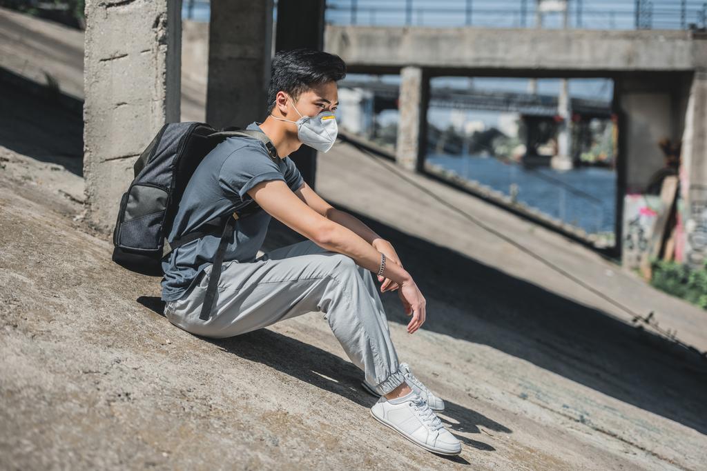 vista lateral de asiático adolescente em máscara protetora sentado sob ponte, conceito de poluição do ar
 - Foto, Imagem