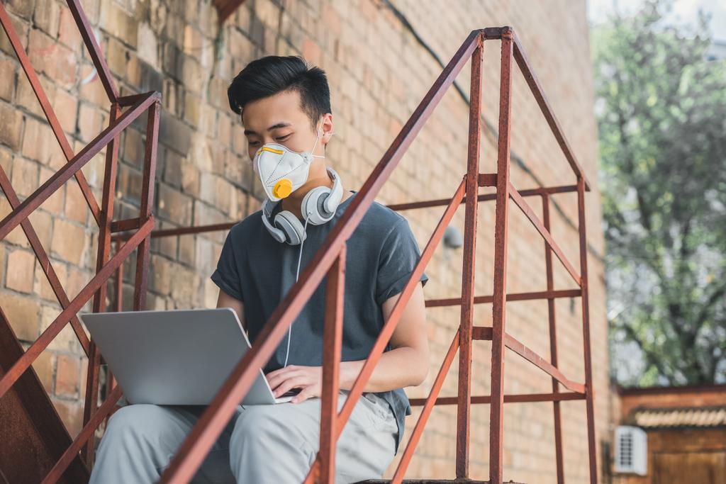 asian teen in protective mask working with laptop on staircase, air pollution concept - Photo, Image