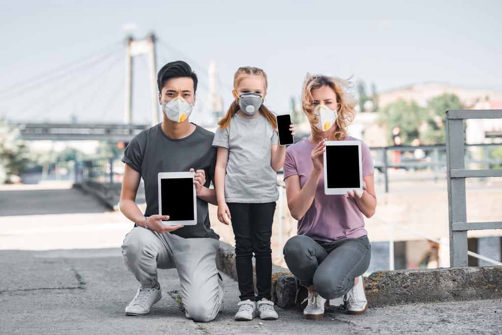 família multicultural com criança em máscaras protetoras segurando gadgets, conceito de poluição do ar
 - Foto, Imagem