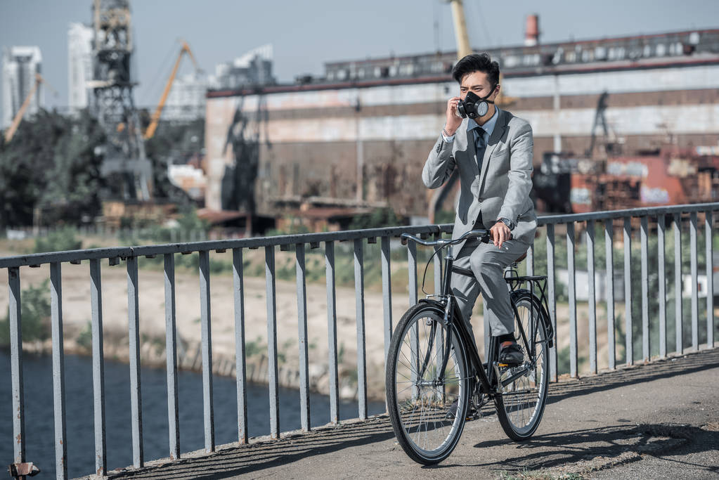 asiatischer Geschäftsmann mit Gasmaske fährt Fahrrad auf Brücke und spricht per Smartphone, Luftverschmutzungskonzept - Foto, Bild