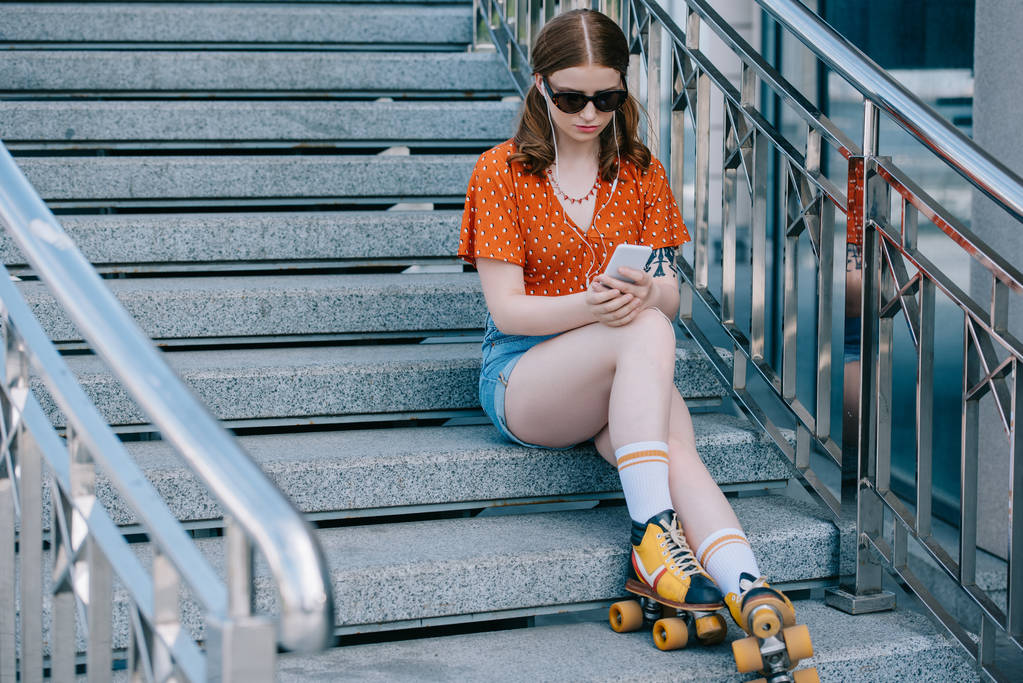 mooi meisje in zonnebrillen en rolschaatsen zittend op trappen en luisteren muziek met smartphone   - Foto, afbeelding