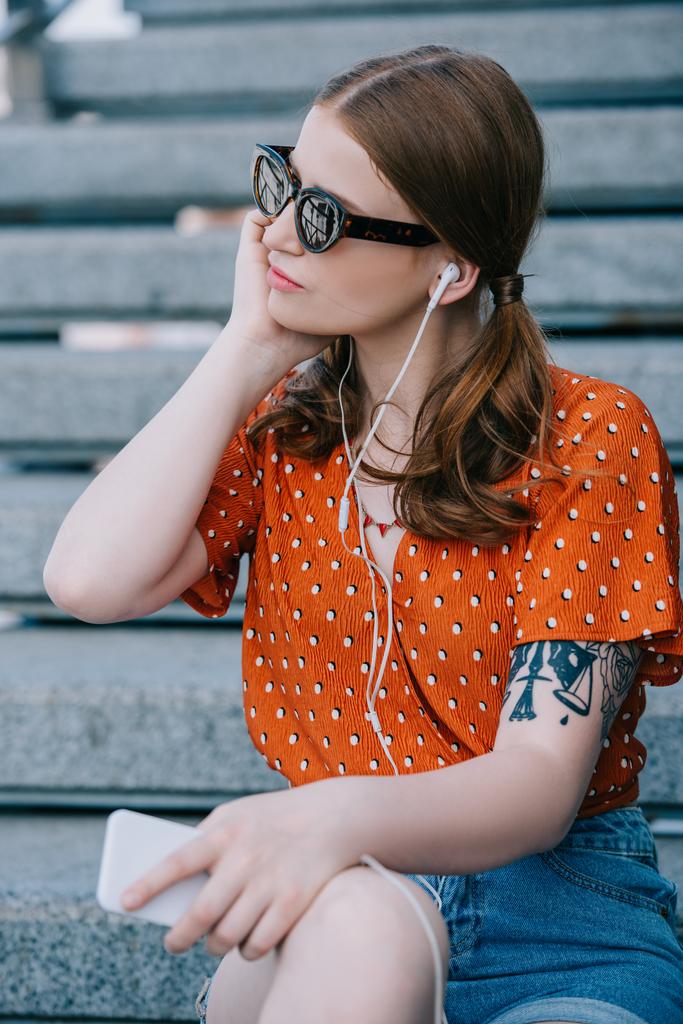 stylová holka v sluneční brýle a sluchátka poslouchat hudbu s smartphone zatímco sedí na schodech - Fotografie, Obrázek