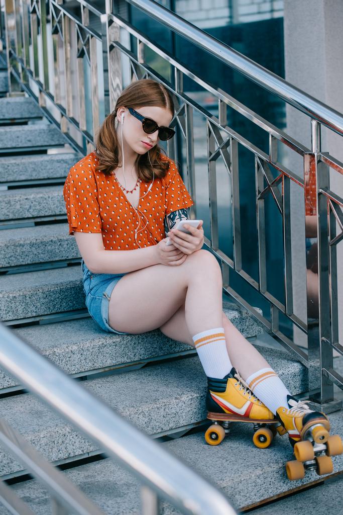 menina elegante em óculos de sol e patins sentados em escadas e ouvir música com smartphone
  - Foto, Imagem