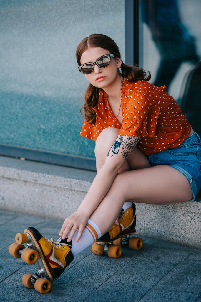meisje in zonnebril camera kijken terwijl zittend in de straat en het dragen van vintage rolschaatsen - Foto, afbeelding