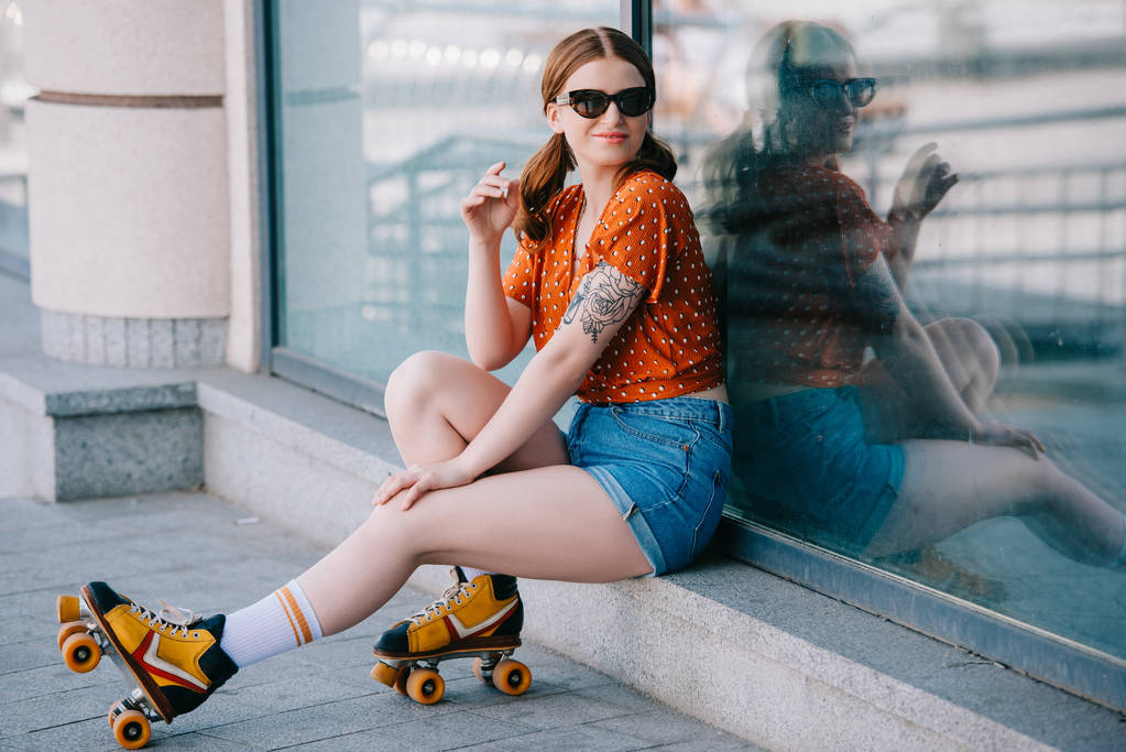 beautiful girl in sunglasses and roller skates smiling at camera while sitting on street - Photo, Image