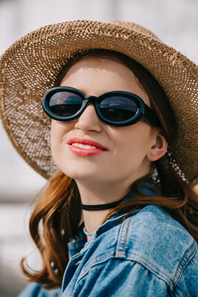 Porträt einer glücklichen jungen Frau mit Sonnenbrille, Hut und Jeansjacke, die draußen lächelt - Foto, Bild