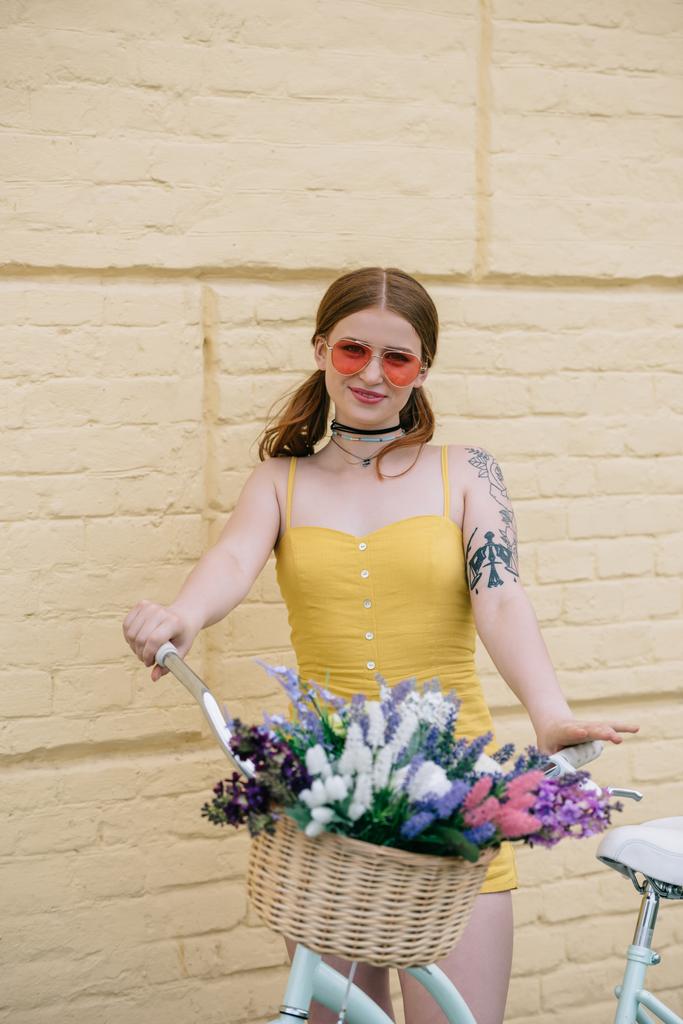 krásná mladá žena v sluneční brýle se usmívá na kameru stoje s kolo na ulici - Fotografie, Obrázek
