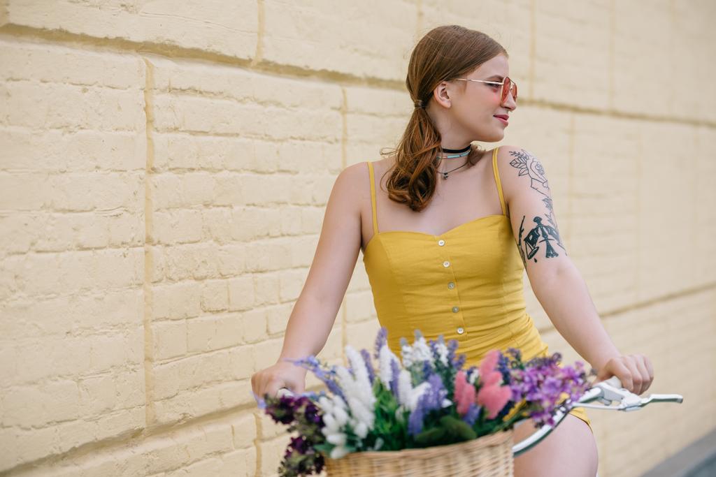 giovane donna sorridente in occhiali da sole in bicicletta con cesto di fiori e guardando lontano sulla strada
 - Foto, immagini