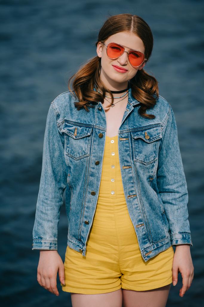 portrait of beautiful young woman in sunglasses and denim jacket looking at camera outdoors - Photo, Image