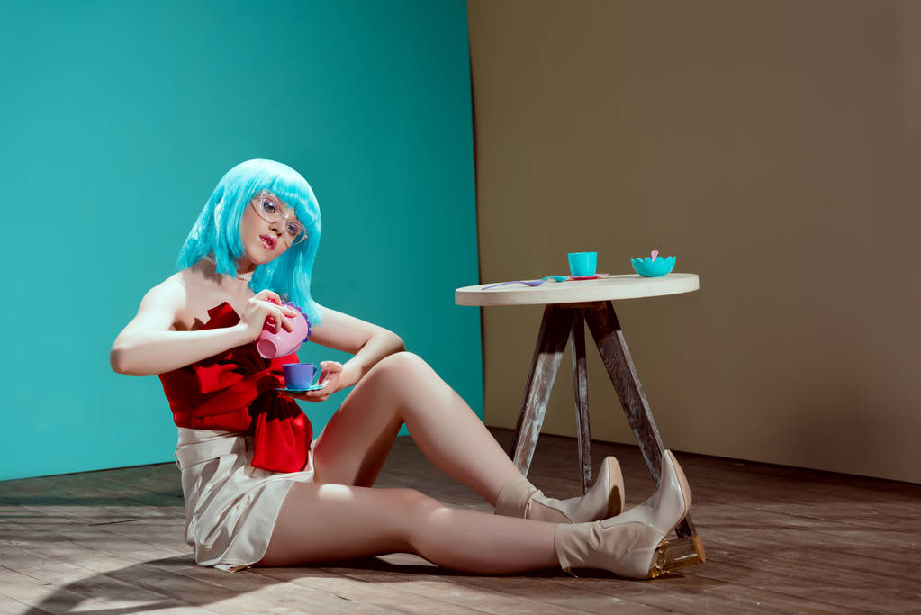 hermosa chica elegante en peluca azul sosteniendo taza de juguete y tetera mientras está sentado en el suelo
  - Foto, Imagen