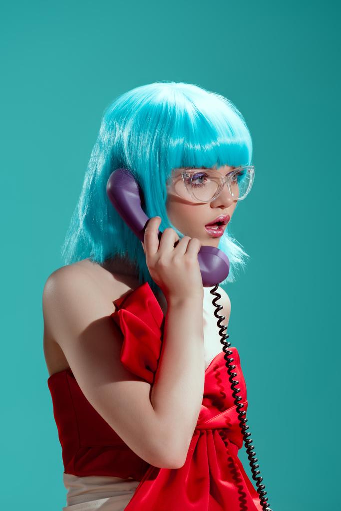 σοκαρισμένος κομψό κορίτσι σε μπλε περούκα μιλώντας τηλεφωνικά και κοιτώντας μακριά απομονωμένη στο μπλε - Φωτογραφία, εικόνα