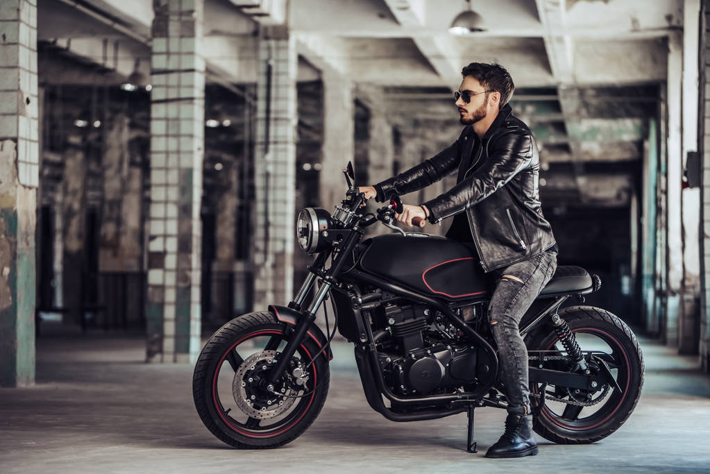 Knappe bebaarde biker met klassieke stijl zwart motorfiets. Cafe racer in garage. - Foto, afbeelding
