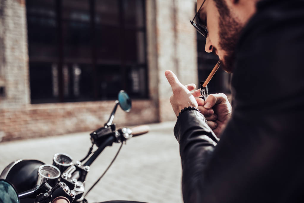 Skadrowana przystojny Brodatego rowerzystę jest palenie sigarette siedząc na styl klasyczny czarny motocykl. Cafe racer. - Zdjęcie, obraz