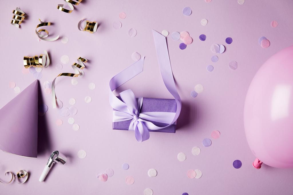 вид зверху на одну фіолетову подарункову коробку, повітряну кулю, капелюх вечірки та фрагменти конфетті на поверхні
 - Фото, зображення