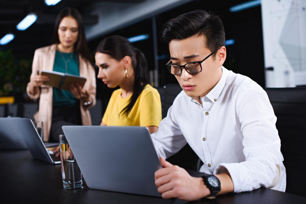 jonge Aziatische zakenman met behulp van laptop terwijl twee vrouwelijke ondernemers werken achter bij moderne kantoor  - Foto, afbeelding
