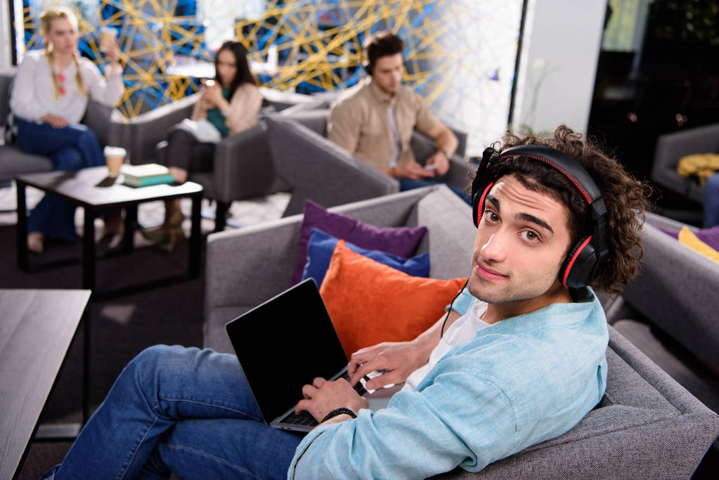 молодий бізнесмен в навушниках сидить на дивані і використовує ноутбук з порожнім екраном в сучасному коворкінгу
  - Фото, зображення