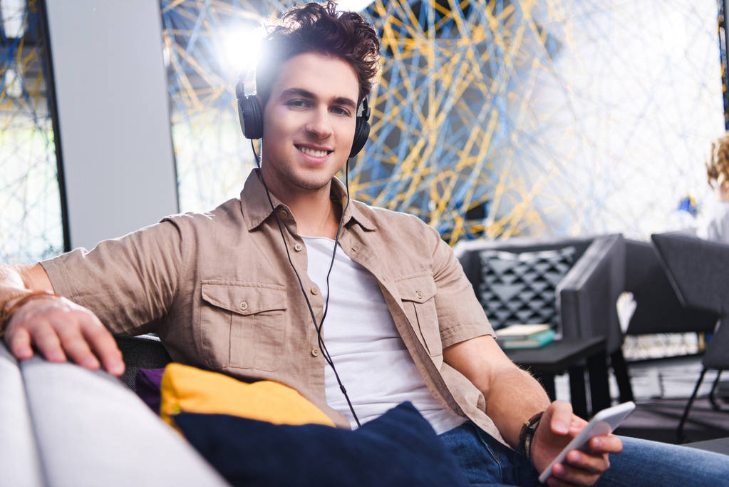 Lächelnder junger Geschäftsmann mit Kopfhörer sitzt mit Smartphone im modernen Coworking-Büro  - Foto, Bild