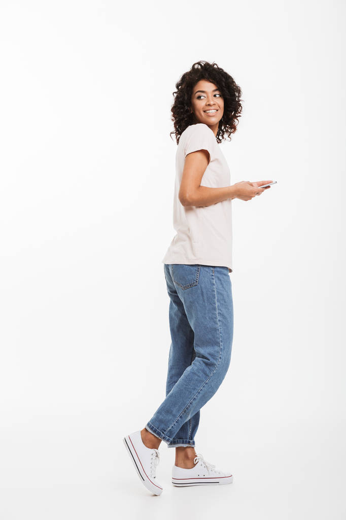 Po celé délce fotografie atraktivní bruneta žena s afro účes nosí tričko a džíny pomocí smartphone a ohlíží se zpět, izolované na bílém pozadí - Fotografie, Obrázek