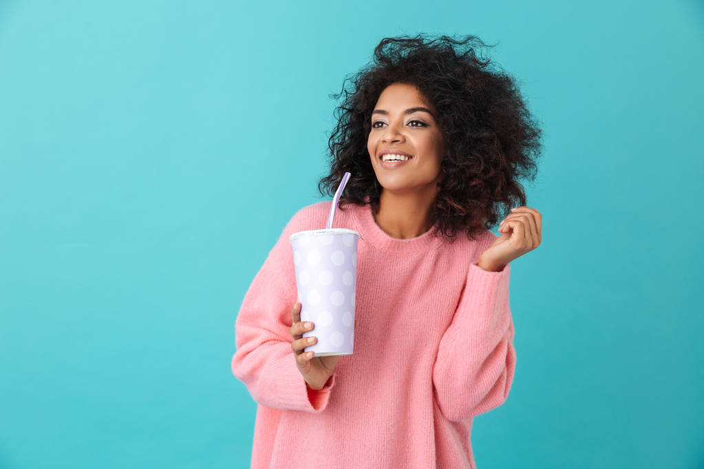 Portrét rozkošné ženy 20s s afro účes Hledáte stranou, zatímco drží nápoj soda v pohárek izolované nad modrým pozadím - Fotografie, Obrázek