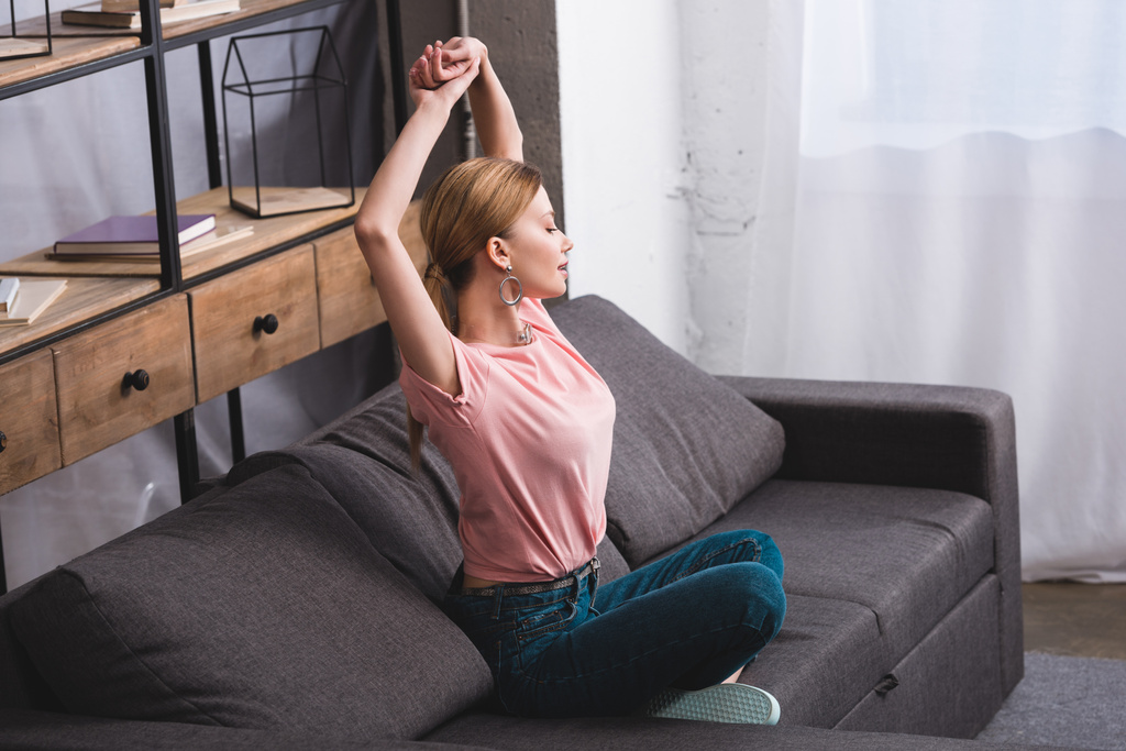 Seitenansicht der attraktiven jungen Frau mit erhobenen Armen, die zu Hause auf dem Sofa sitzt - Foto, Bild