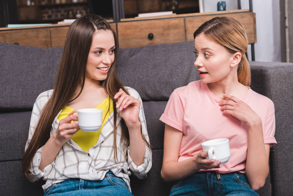 две юные подруги с чашками кофе сидят дома на диване
 - Фото, изображение