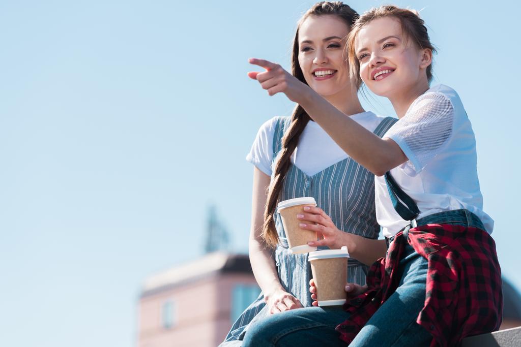 Genç kadın kağıt bardak kahve ile gülümseyen kadın arkadaşına parmak tarafından işaret  - Fotoğraf, Görsel