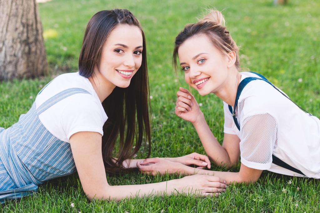 junge lächelnde Freundinnen liegen auf grünem Rasen im Park  - Foto, Bild