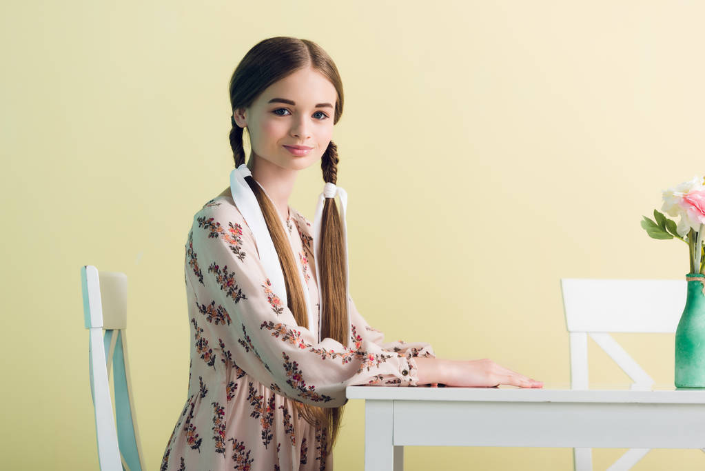 adolescente en vestido de verano con trenzas sentado a la mesa con flores, aislado en amarillo
 - Foto, imagen