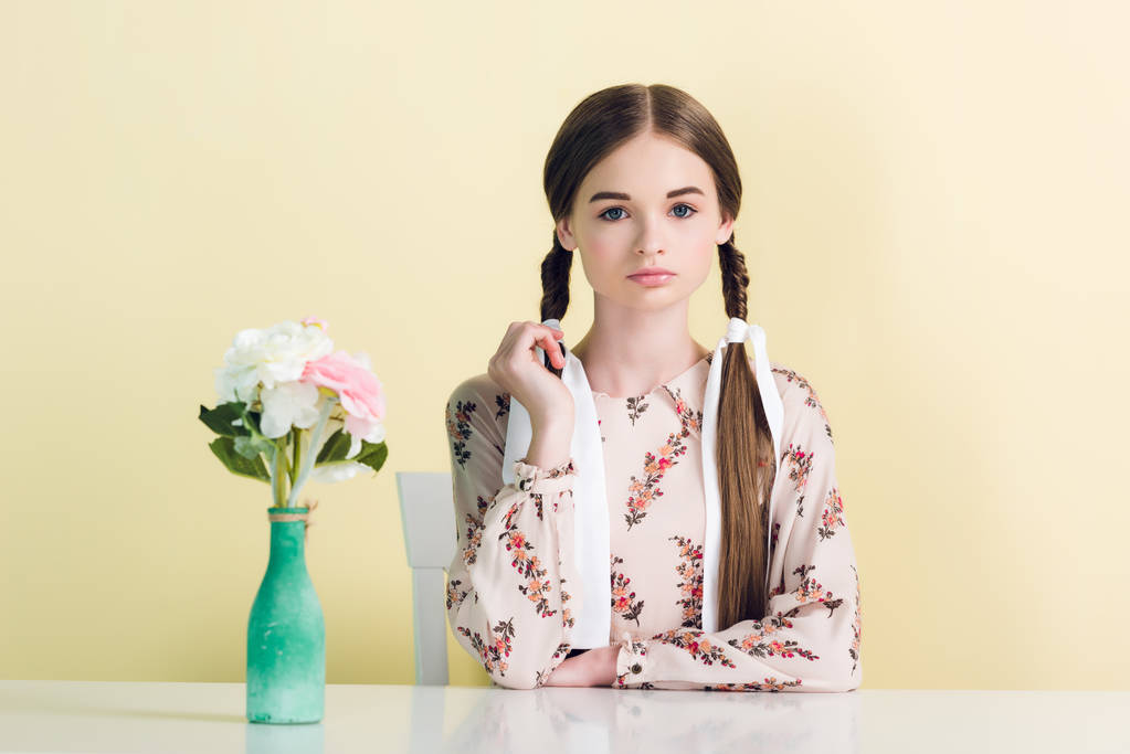 elegante ragazza adolescente con trecce seduto a tavola con vaso e fiori, isolato su giallo
 - Foto, immagini