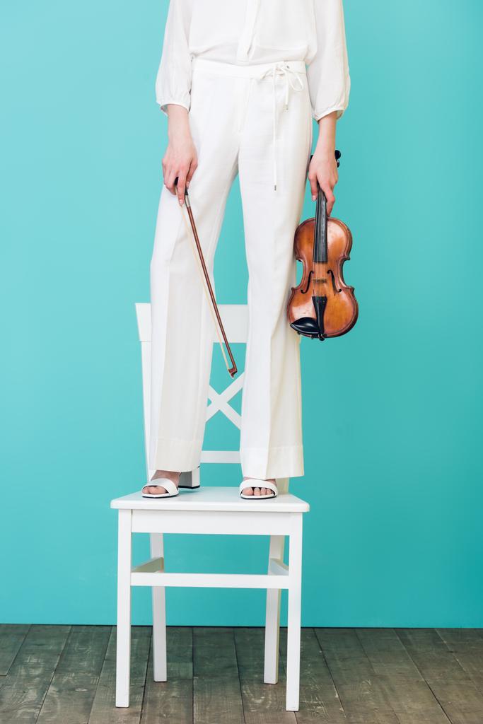 Обрезанный вид девочки, держащей скрипку и стоящей на стуле, на голубом
 - Фото, изображение