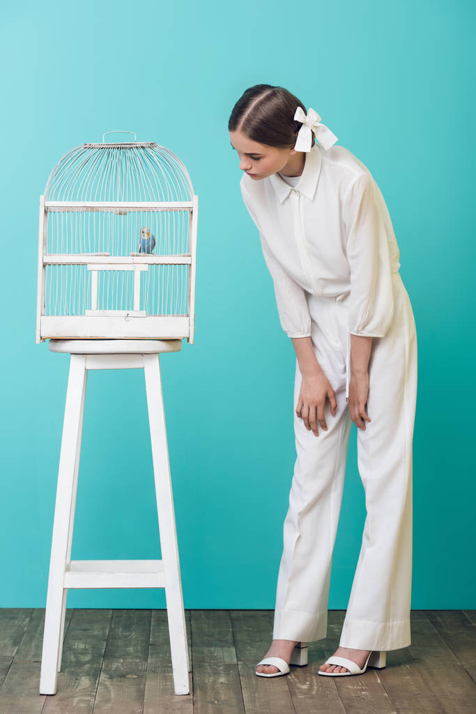 attractive girl in white looking at parrot in cage, on turquoise - Photo, Image