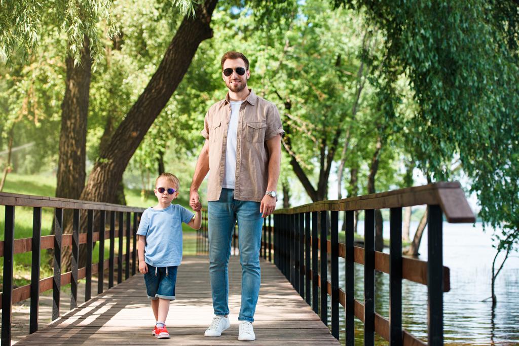 батько і син в сонцезахисних окулярах тримаються за руки і ходять по мосту в парку
 - Фото, зображення