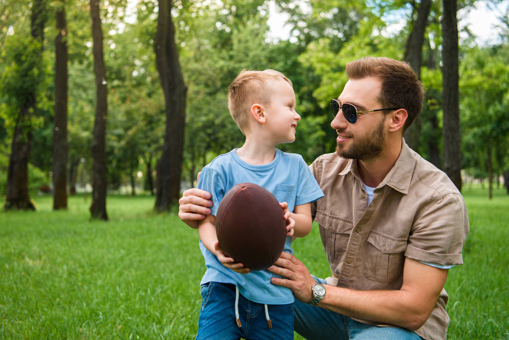 ευτυχής πατέρας και γιος βλέπουν ο ένας τον άλλον και κρατώντας αμερικανικό ποδόσφαιρο μπάλα στο πάρκο - Φωτογραφία, εικόνα