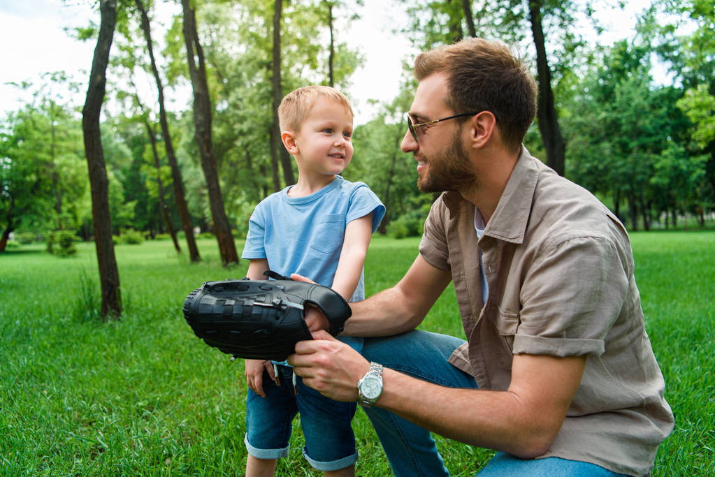 father wearing baseball glove on son hand at park - Photo, Image