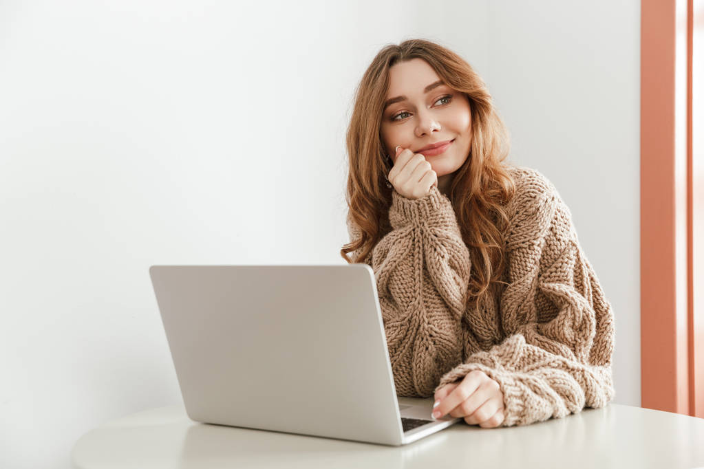 Imagem de mulher bonita 20s em suéter com cabelos castanhos longos olhando para o lado e usando laptop enquanto sentado à mesa no restaurante
 - Foto, Imagem