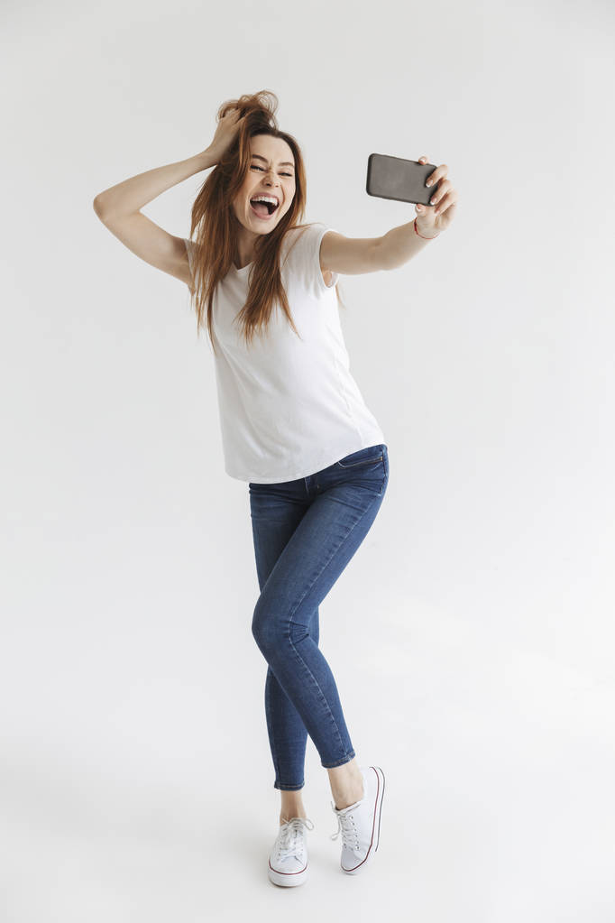 Volledige lengte beeld van blijde vrouw in casual kleding poseren en het maken van de selfie op smartphone over grijze achtergrond - Foto, afbeelding
