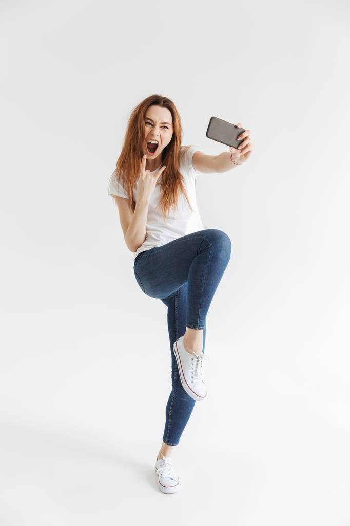 Pełna długość obrazu kobiety Screaming w ubranie podejmowania selfie na smartphone i pokazuje gest rock na szarym tle - Zdjęcie, obraz