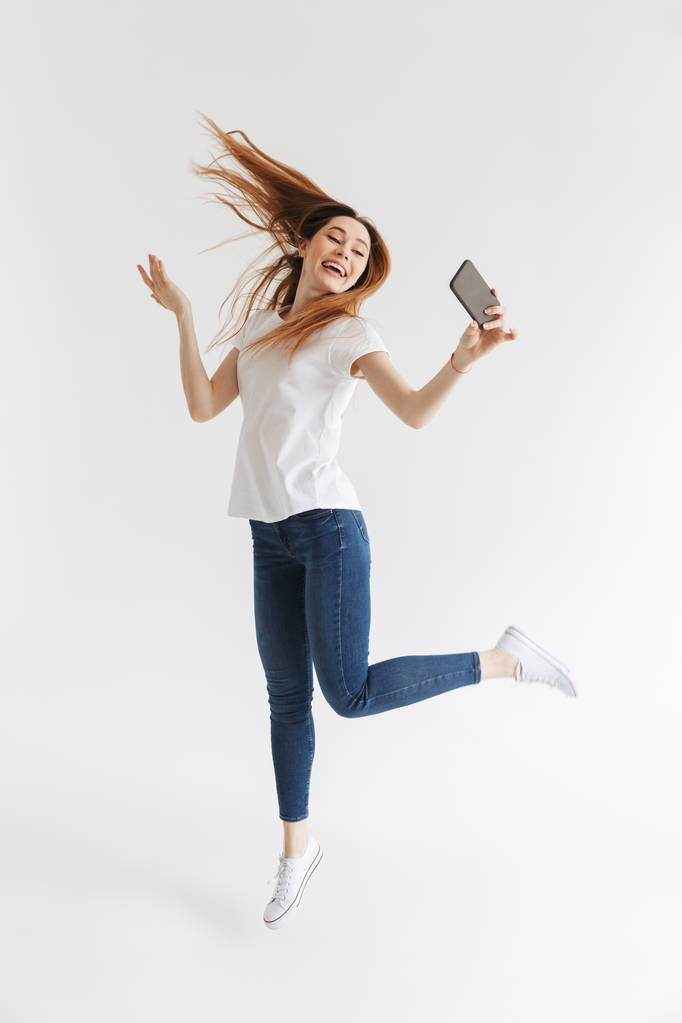 Tam boy görüntü atlama ve selfie smartphone üzerinde gri arka plan üzerinde yapma rahat kıyafetler içinde neşeli kadın - Fotoğraf, Görsel