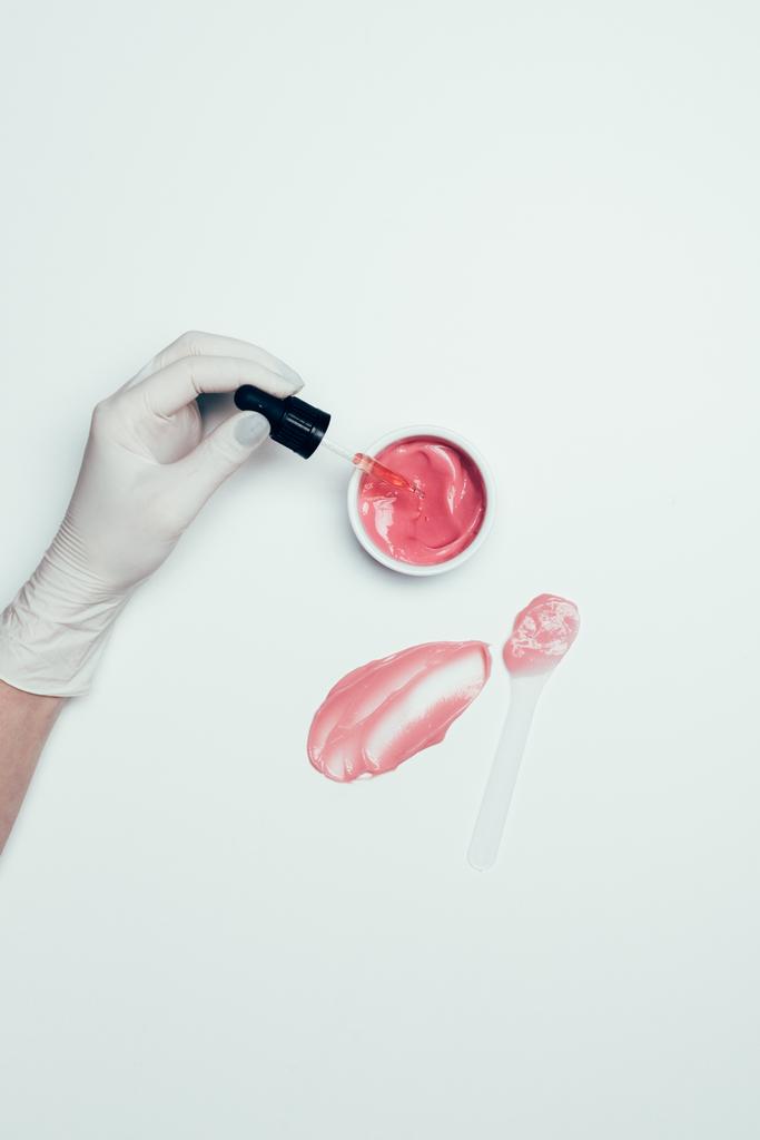 Uma mão em luva de látex segurando tubo perto do recipiente com máscara rosa, colher e mancha na superfície branca
 - Foto, Imagem