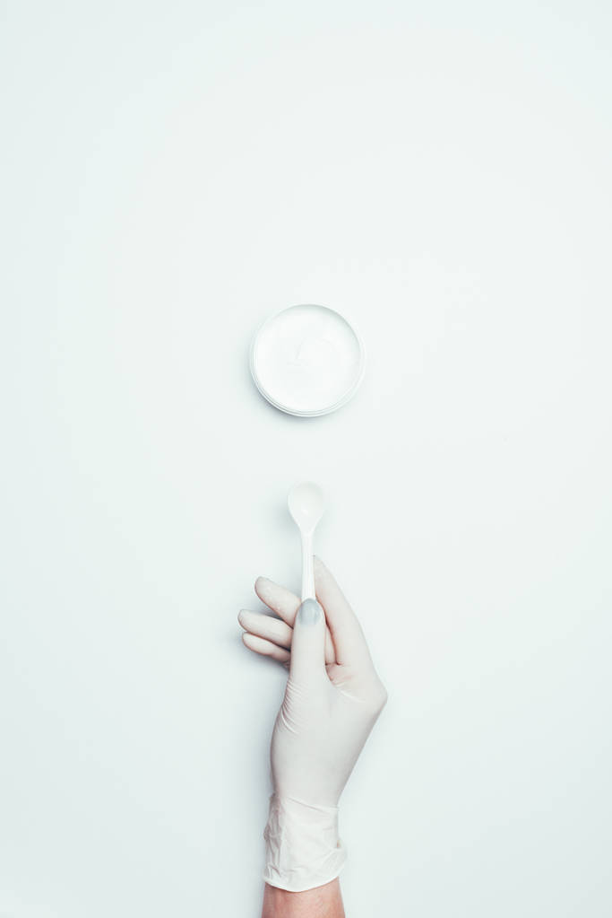 περικομμένη εικόνα της γυναίκας στην latex γάντι κρατά κουτάλι κοντά σε δοχείο με μάσκα αργίλου που απομονώνονται σε λευκή επιφάνεια  - Φωτογραφία, εικόνα