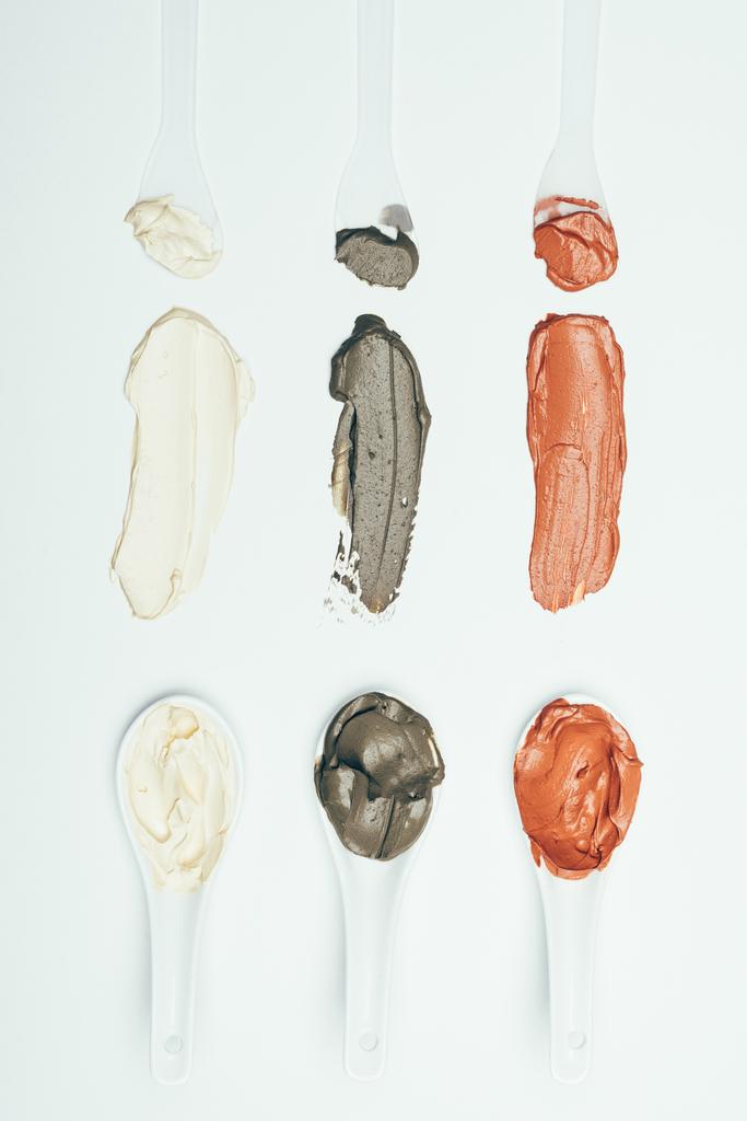 flache Lage mit Löffeln und verschiedenen bunten Maskenflecken aus Ton, die in einer Reihe auf weißer Oberfläche isoliert platziert sind  - Foto, Bild
