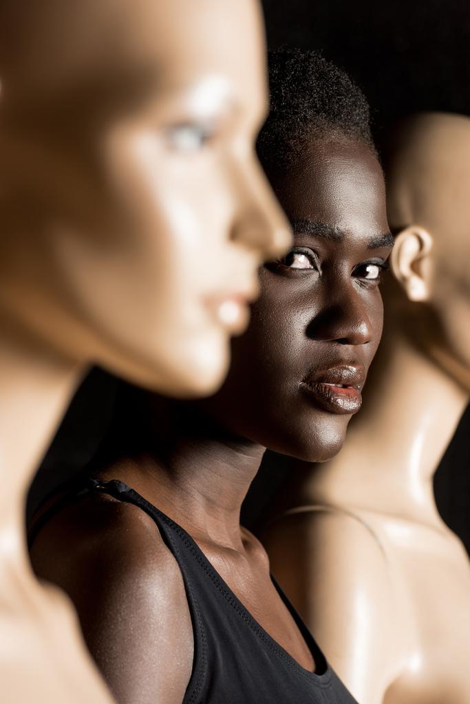 selectieve aandacht van prachtige Afrikaanse Amerikaanse vrouw kijken camera terwijl staande tussen dummies op zwart  - Foto, afbeelding